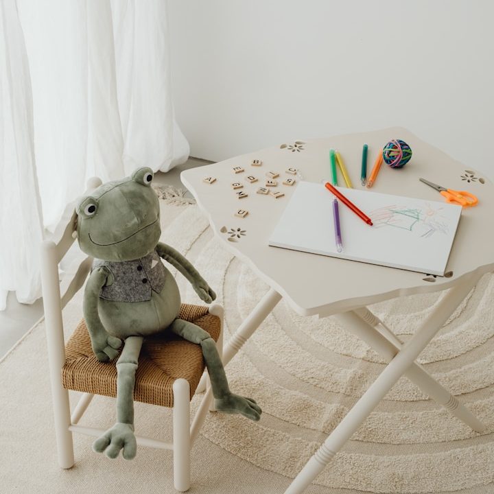 Therapie für Kinder bei Göppingen | Praxis Rauschmaier (häufig gesucht als Kinderpsychologe) Ein grüner Plüschfrosch sitzt auf einem Stuhl neben einem Tisch mit Bastelmaterialien.